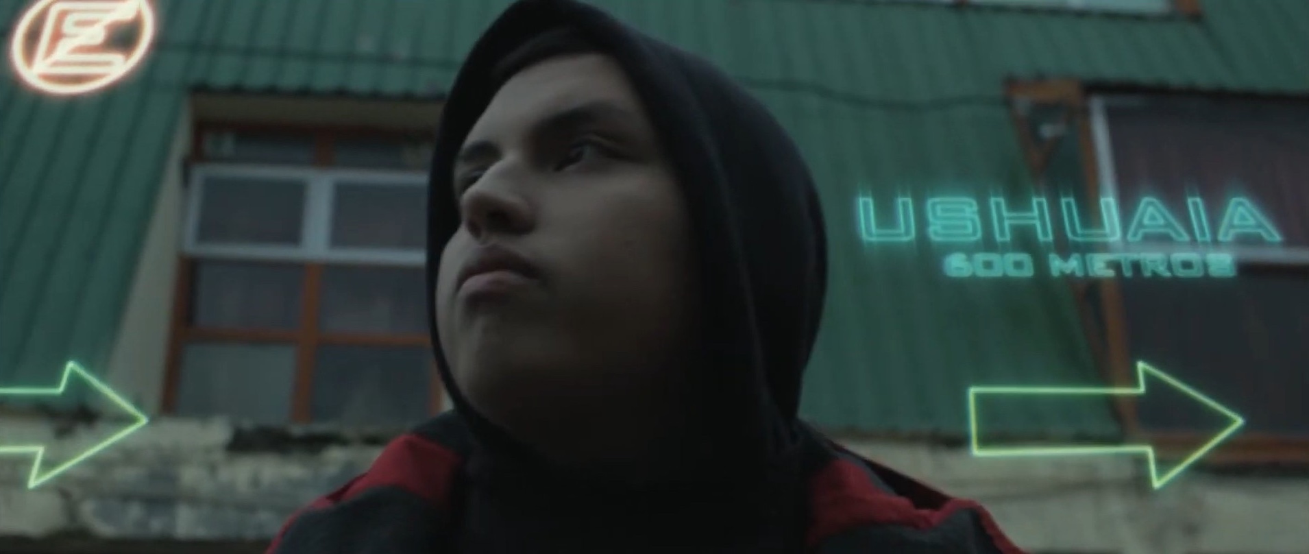 Joven con capucha en contrapicado frente a un cartel de neón: Ushuaia 600 metros